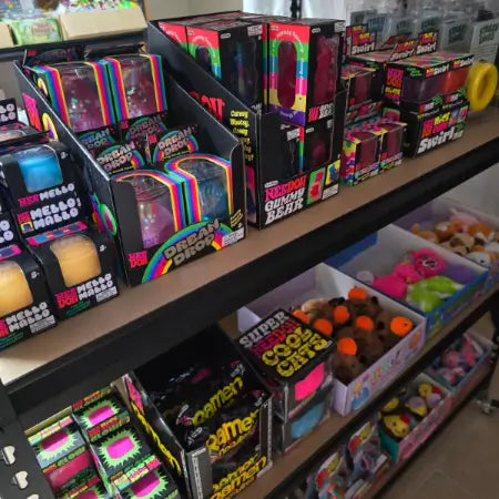 Shelves stocked with colorful novelty toy and accessory boxes in a retail store setting. Visible products include rainbow-striped packaged items, black boxes with neon text, and bins of plush toys and small accessories-fun fidgets