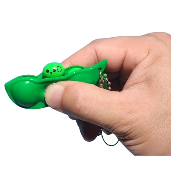Hand holding a green pea pod keychain with a small green character inside on a white background